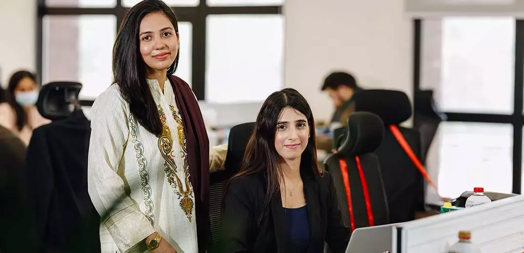 Two women in an office setting, one standing beside the other, who is seated at a workstation with a computer.