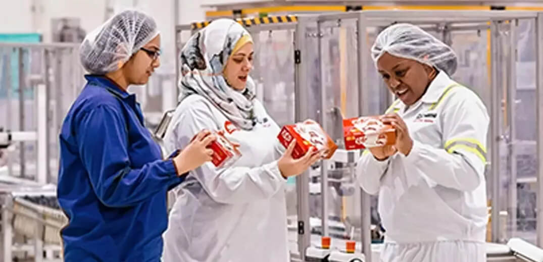 Three individuals in a food processing facility hold boxes, engaged in conversation while wearing protective clothing.