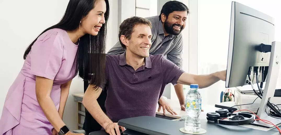A group of three colleagues engage collaboratively around a computer, with a water bottle and headphones on the desk.