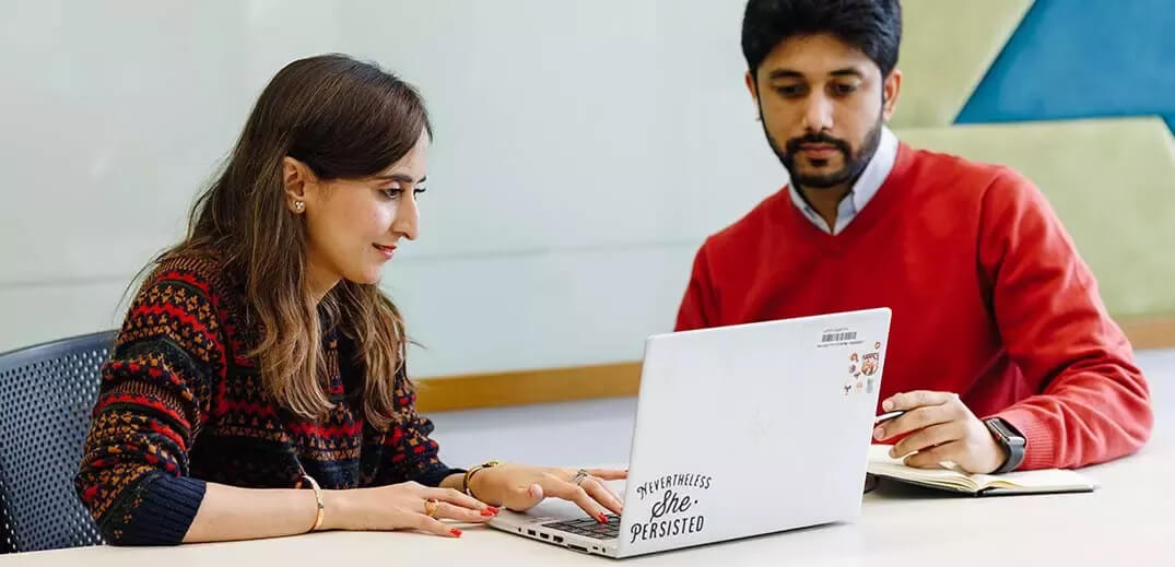 A woman in a patterned sweater and a man in a red sweater discuss a laptop on a table, surrounded by a modern workspace.