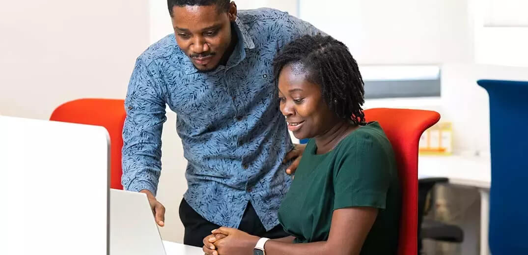 Two people are engaged in a work discussion, leaning over a laptop in a modern office setting with red and blue chairs.