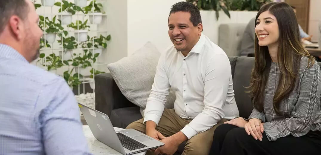 A laptop sits on a table as two people sit on a couch, engaged in conversation in a modern, plant-filled office space.