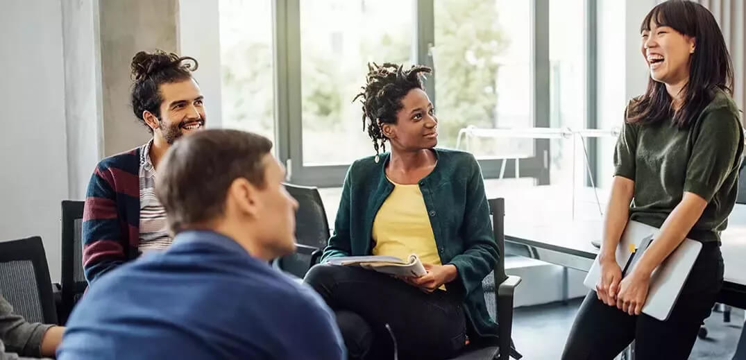A diverse group of individuals engaged in a discussion in a brightly lit, modern meeting room with large windows.