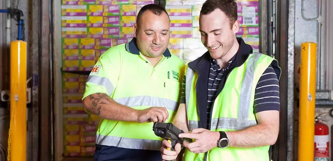 Two warehouse workers, wearing reflective vests, check a handheld scanner against colorful stacked boxes in the background.
