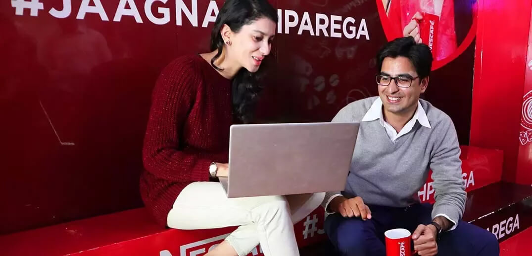 A woman in a maroon sweater and a man in a grey sweater sit together, focused on a laptop in a vibrant, branded setting.