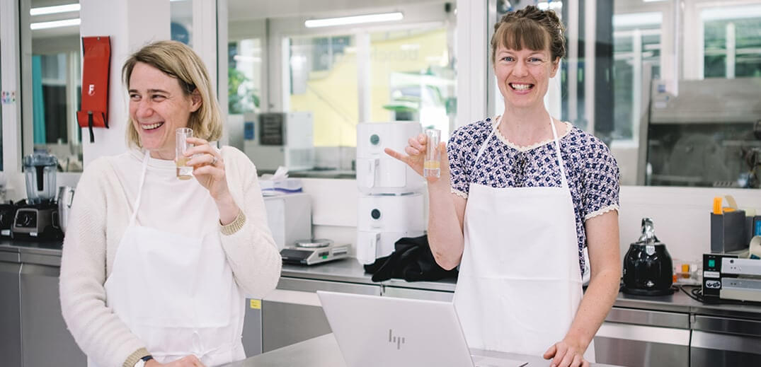 Two women in aprons hold small glass containers in a bright kitchen with modern appliances and a laptop on the counter.