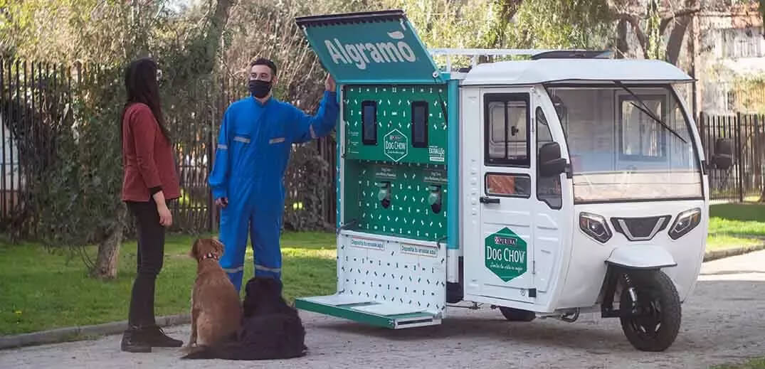 A woman interacts with a man by a pet food distribution vehicle, with two dogs sitting nearby in a park setting.
