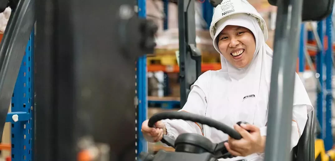 A person in a white uniform and a hat operates a forklift in a warehouse, surrounded by shelves filled with goods.