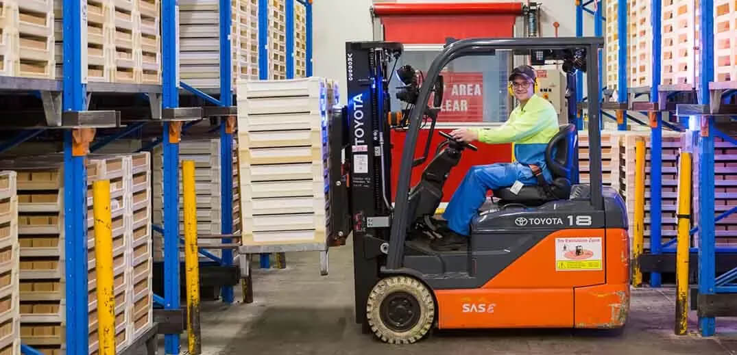 A forklift operator maneuvers a forklift, lifting a stack of wooden pallets in a warehouse aisle lined with storage racks.