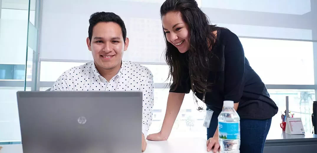 A man and a woman collaborate at a desk, focused on a laptop, with a water bottle and office supplies nearby.