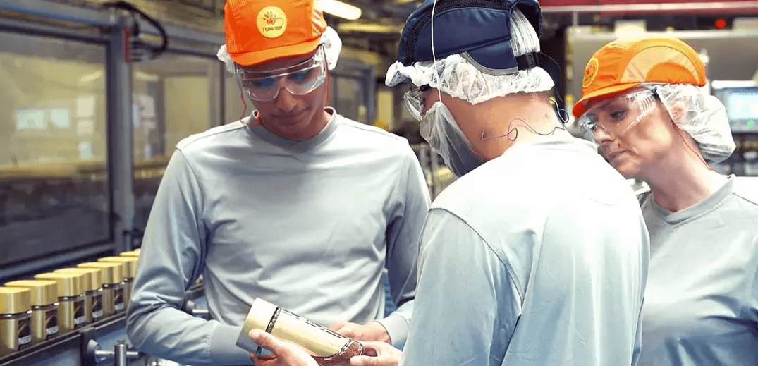 Three factory workers discuss a product while surrounded by jars on a production line, wearing safety gear and hairnets.